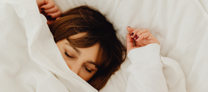 Mulher dormindo confortavelmente em cama com roupa de cama branca, transmitindo sensação de maciez e aconchego.